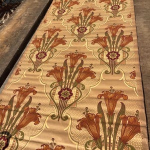 Puede incluir: Un rollo de papel tapiz vintage con un patr&oacute;n repetido de lirios naranjas, hojas verdes y dise&ntilde;os geom&eacute;tricos marrones y verdes sobre un fondo beige.