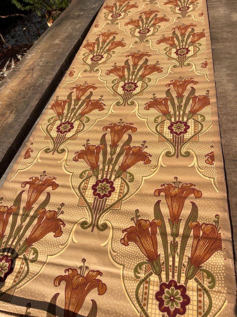 Puede incluir: Un rollo de papel tapiz vintage con un patr&oacute;n repetido de lirios naranjas, hojas verdes y dise&ntilde;os geom&eacute;tricos marrones y verdes sobre un fondo beige.