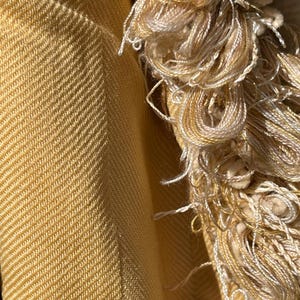 May include: Close-up of a yellow woven fabric with a fringe border. The fabric has a subtle herringbone pattern and a soft, textured feel.
