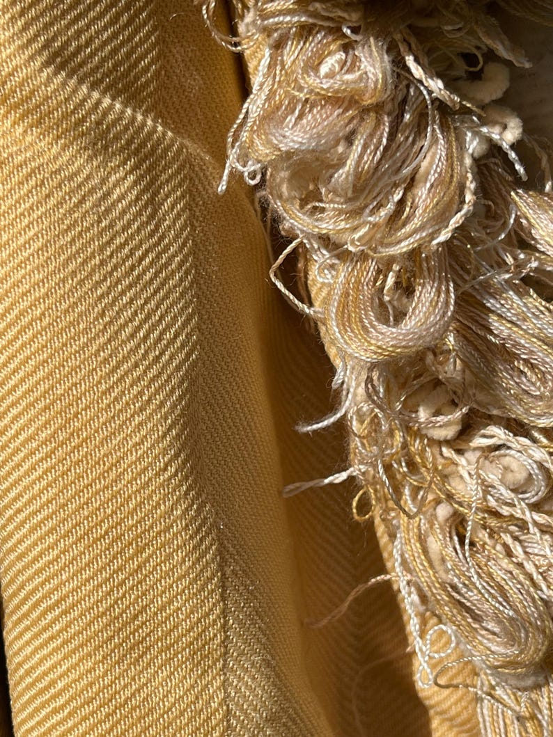 May include: Close-up of a yellow woven fabric with a fringe border. The fabric has a subtle herringbone pattern and a soft, textured feel.