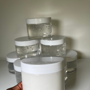 Peut inclure: Une pile de pots blancs contenant un gel transparent, avec un pot blanc au premier plan contenant une crème blanche.