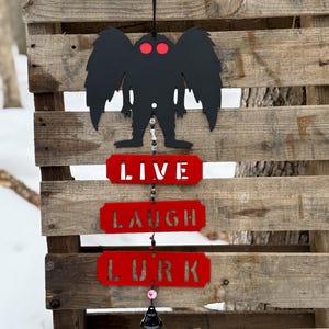 May include: Black metal wall hanging featuring a silhouette of a winged creature with red eyes, above three red rectangular signs that read "LIVE", "LAUGH", and "LURK" in white letters. The hanging is suspended from a black string.