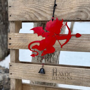 May include: A red metal silhouette of a devilish Cupid with a bow and arrow, wings, and a tail, hanging from a black cord. A small black bell and beads dangle below. The design is set against a wooden pallet.