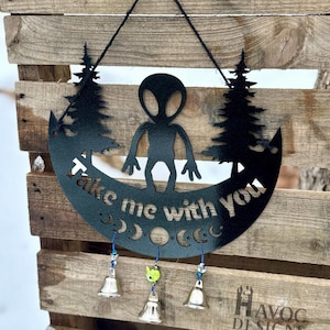 May include: Black metal wall hanging featuring an alien figure, trees, and the phrase "Take me with you." The design includes crescent moon shapes and three small bells. The piece is suspended by a black cord and is mounted on a wooden pallet.