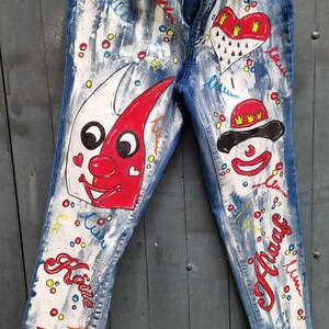 May include: Blue denim jeans with white paint splatters and colorful hand-painted designs. The designs include a clown face, a heart with a crown, and the word "Katie".