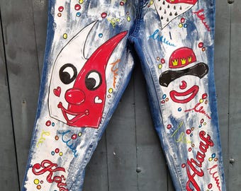 May include: Blue denim jeans with white paint splatters and colorful hand-painted designs. The designs include a clown face, a heart with a crown, and the word "Katie".