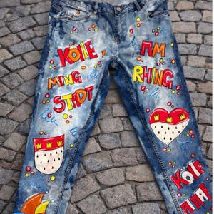 May include: Blue denim jeans with colorful hand-painted designs. The jeans feature text and illustrations in red, yellow, and orange, including the words "KÖLLE", "MING", "STADT", and "AM RHING". Decorative elements include hearts and a carnival hat.