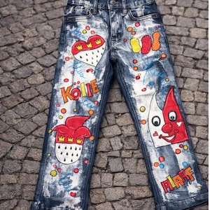 May include: Blue denim jeans with colorful painted designs. Features include a heart-shaped emblem with a crown, a jester's hat, and the word "KOLLE" in orange. The jeans are decorated with various shapes and colors.