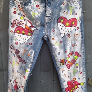 May include: Blue denim jeans with white paint splatters and colorful hand-painted designs. The designs include hearts, crowns, and the words "Kölle Alaaf!"