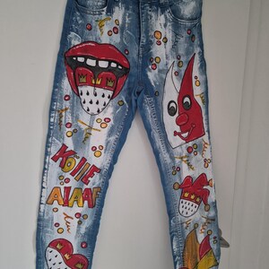May include: Blue denim jeans with hand-painted designs. The jeans feature a red mouth, a jester, and the text "KOLIE ALAAF" in white. The jeans are decorated with colorful accents and a heart.
