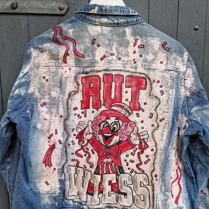 May include: Blue denim jacket with a colorful clown graphic on the back. The artwork features the words "RUT" and "WIESS" in bold letters, along with red confetti and accents. The jacket is hanging on a white hanger.