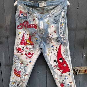 Könnte beinhalten: Hellblaue Jeans aus Denim mit handgemalten Designs. Die Jeans zeigen zwei Clowns, ein rotes und weißes Gesicht und den Text "Alaar", "E Leyve Lang!" und "Kolsch".
