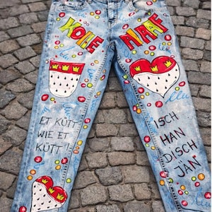 May include: Light blue denim jeans with colorful hand-painted designs. The jeans feature the words "KOLE" and "HAF" in yellow and red, along with heart and crown motifs. Additional text and colorful dots are scattered across the jeans.