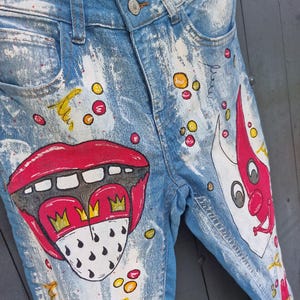 May include: Light blue slim fit denim jeans with colorful hand-painted artwork. The design includes a large red mouth with a crown, and other abstract shapes. The jeans have a distressed look with white paint splatters.