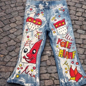 May include: Light blue denim jeans with colorful hand-painted designs. Features a heart with a crown, a red and white clown face, and the text "KÖLE HAAAF". The jeans have a flared leg opening.