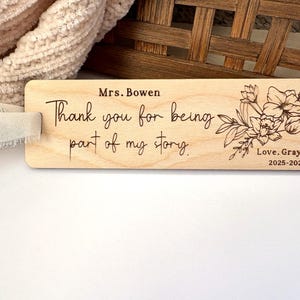 May include: A rectangular wooden bookmark with the text "Mrs. Bowen, Thank you for being part of my story. Love, Grayson 2025-2026" and a floral design. The bookmark is light brown with dark brown engraved text and a floral design.