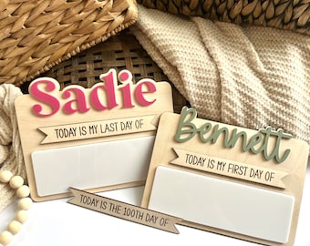 May include: Two wooden signs with blank spaces for writing. The signs are decorated with the names "Sadie" and "Bennett" in pink and green letters. The signs also have banners that say "Today is my last day of" and "Today is my first day of".