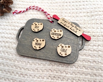 May include: Wooden Christmas ornament shaped like a baking sheet with four cookie cutouts. Each cookie is personalized with a name: Rex, Grayson, Noah, and Cole. A red rolling pin with the text "Gigi & Pop Pop's perfect batch" is attached to the baking sheet.