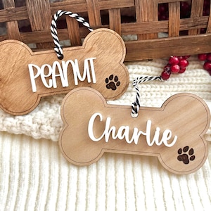 May include: Two wooden dog bone ornaments with black and white twine hangers. The ornaments are personalized with the names "Peanut" and "Charlie" in white lettering. Each ornament has a paw print design.