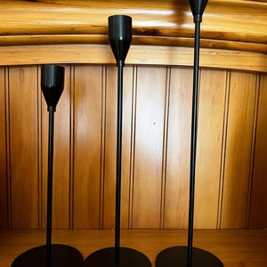 May include: Three black metal candle holders of varying heights. Each holder has a circular base, a slender stem, and a cup-shaped top designed to hold a candle. The holders are arranged in a row, showcasing their minimalist design.