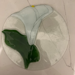 May include: A white glass plate with a green and white flower design. The flower is a calla lily with a yellow center.