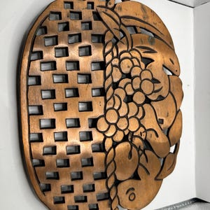 May include: An oval wooden trivet featuring a carved design of grapes and leaves on one side and a geometric basket-weave pattern on the other. The natural light brown wood has dark brown accents within the carved details and cut-out sections.