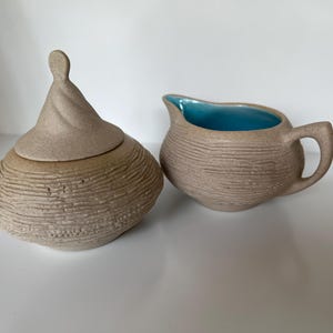 May include: A set of two ceramic sugar and creamer containers. The sugar bowl has a lid with a unique, textured design. The creamer has a light blue interior and a textured exterior.