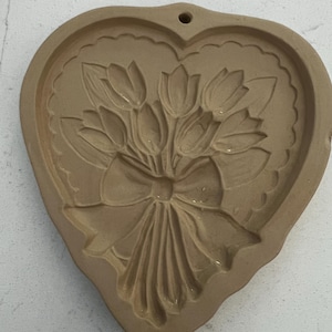 May include: A heart-shaped, tan-colored mold with a floral design. The mold features a bouquet of tulips tied with a bow. The edges of the heart have a scalloped design, and there is a hole at the top for hanging.