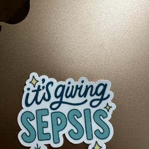 Puede incluir: Una pegatina blanca con la frase "it's giving SEPSIS" en letras turquesas y azul oscuro. La pegatina tiene un borde blanco y detalles decorativos de estrellas y copos de nieve. La pegatina est&aacute; sobre una superficie marr&oacute;n.
