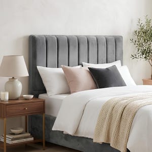 May include: A grey velvet upholstered headboard with vertical channel tufting. The bed is dressed with white linens, a beige knit throw blanket, two white pillows, one light pink pillow, and one dark grey pillow, all arranged against the headboard.