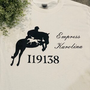 May include: White t-shirt with a black silhouette of a person riding a horse jumping over a fence. The text "Empress Karolina" and the number "119138" are printed below the image.