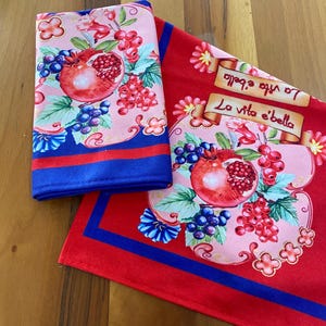 May include: Two red kitchen towels with a blue border. The towels feature a pomegranate, grapes, and flowers. One towel has the text "La vita e' bella" and "Io amo la bella" in a scroll.