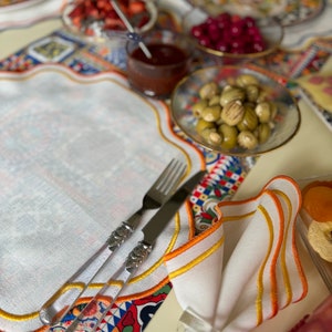 Puede incluir: Una mesa decorada con un mantel individual blanco con borde festoneado, una servilleta blanca doblada con ribete naranja y amarillo, y cubiertos con mango transparente. Se ven pequeños cuencos con aceitunas, fresas y cerezas, junto con un vaso pequeño de salsa. La mesa está decorada con manteles estampados coloridos.