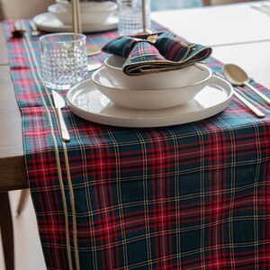 May include: A dining table set with a red, green, and gold tartan table runner. White plates and bowls are stacked, with a tartan napkin on top. Gold cutlery and glassware complete the festive arrangement.