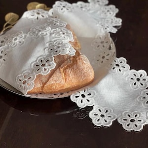 Peut inclure: Une miche de pain sur une assiette argentée, partiellement recouverte d'une serviette en lin blanche. La serviette présente un motif floral ajouré délicat sur les bords. Le pain a une croûte brun doré.