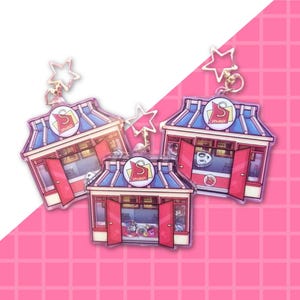 May include: Three acrylic keychains shaped like small shops with red doors, blue roofs, and a circular sign featuring the letter S. Each keychain includes a gold-coloured star-shaped clasp attached to the top of the structure.