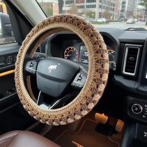 May include: A crocheted steering wheel cover in shades of brown and tan, installed on a car's steering wheel. The cover features a textured pattern and adds a decorative element to the vehicle's interior.