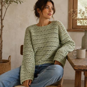May include: A sage green long-sleeve crochet sweater featuring a repeating pattern of solid horizontal bands and openwork grid sections, a round neckline, and loose-fitting sleeves, paired with light blue denim jeans and white sneakers.