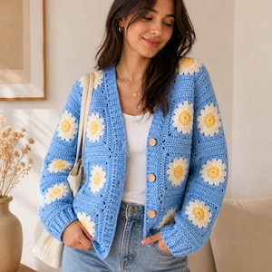 May include: A long-sleeve crochet cardigan in light blue featuring a repeating pattern of white and yellow daisy motifs. The garment has a V-neckline, a button-front closure with four round wooden buttons, and ribbed cuffs at the wrists.