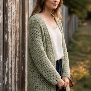 May include: A light green, open-weave crochet cardigan. The cardigan is worn over a white top and dark blue jeans. The model is standing next to a weathered wooden fence, with a brown purse.