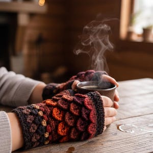 May include: A person holding a steaming brown mug, wearing crochet fingerless gloves. The gloves feature a scale-like pattern in shades of brown, orange, and red. The scene is set on a wooden table, with a blurred background of a cozy interior.