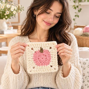 Könnte beinhalten: Ein quadratisches Häkelmotiv mit einem zentralen rosa Apfeldesign und einem kleinen braunen Stiel, umrandet von einer cremefarbenen Bordüre. Das Stück ist mit einem strukturierten Garnstich gefertigt, der ein geometrisches Muster mit offenen Ecken bildet.