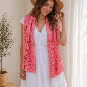 May include: A woman wearing a straw hat and a coral-colored, open-weave crochet shawl over a white dress. The shawl has a scalloped edge. A small plant in a woven basket sits on a wooden table.