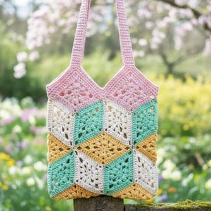 May include: A handmade crochet tote bag with a geometric pattern. The bag features a pink top, with a body of alternating cream, teal, and yellow diamond shapes. The bag is suspended in front of a blurred background of flowers and greenery.