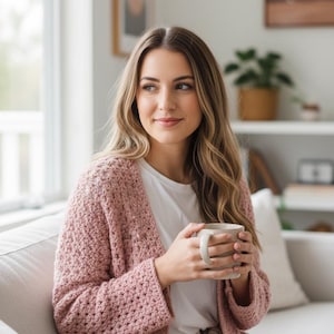 Puede incluir: Una persona con un cárdigan de ganchillo rosa claro sobre una camiseta blanca, sosteniendo una taza beige. El cárdigan tiene un patrón de puntada texturizada. La escena es en interiores con luz natural.