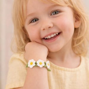 May include: A smiling child wearing a yellow shirt and a crochet daisy bracelet. The bracelet features white daisy flowers with yellow centers and green leaves. The child has blonde hair and is resting their chin on their hand.