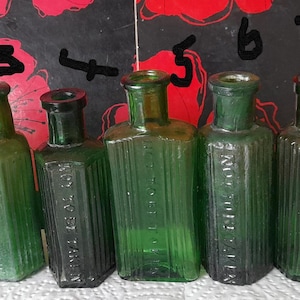 May include: A selection of nine antique green glass bottles in various shapes and sizes. Some bottles feature embossed text, all with a vintage appearance. The bottles are displayed in a row, highlighting their diverse designs and heights.
