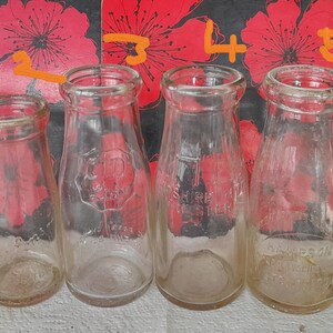 Può includere: Sei bottiglie di latte vintage in vetro trasparente di varie dimensioni. Le bottiglie hanno una forma arrotondata con colli stretti e scritte in rilievo. Alcune bottiglie hanno testo visibile, tra cui "ROSE DAIRY" e "CO".