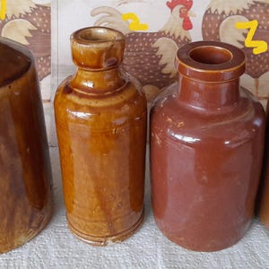 Puede incluir: Cuatro botellas antiguas de cerámica marrón de diferentes formas y tamaños. Las botellas tienen un acabado brillante y se exhiben sobre una superficie blanca. El fondo presenta una ilustración de gallo.