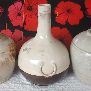Może przedstawiać: Trzy antyczne wazy ceramiczne o różnych kształtach i kolorach. Wazy są w kolorze ecru, z brązowymi i szarymi akcentami. Tło przedstawia czarno-czerwony wzór kwiatowy.
